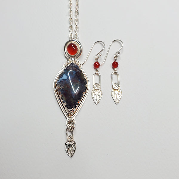 A handmade argentium silver kite shaped pendant featuring a rich green, blood red and white moss agate stone, The carnelian accent cabochon bezel set above symbolizes the energy that a seed needs in the process of germination. The details are hand stamped and formed using handmade stamps from the Navajo Nation. The accompanying earring contain carnelian beads.